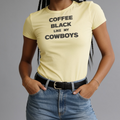 A woman wearing a pale yellow graphic t-shirt with the text 'COFFEE BLACK LIKE MY COWBOYS' in black capital letters, paired with blue jeans and a black belt, against a solid gray background.